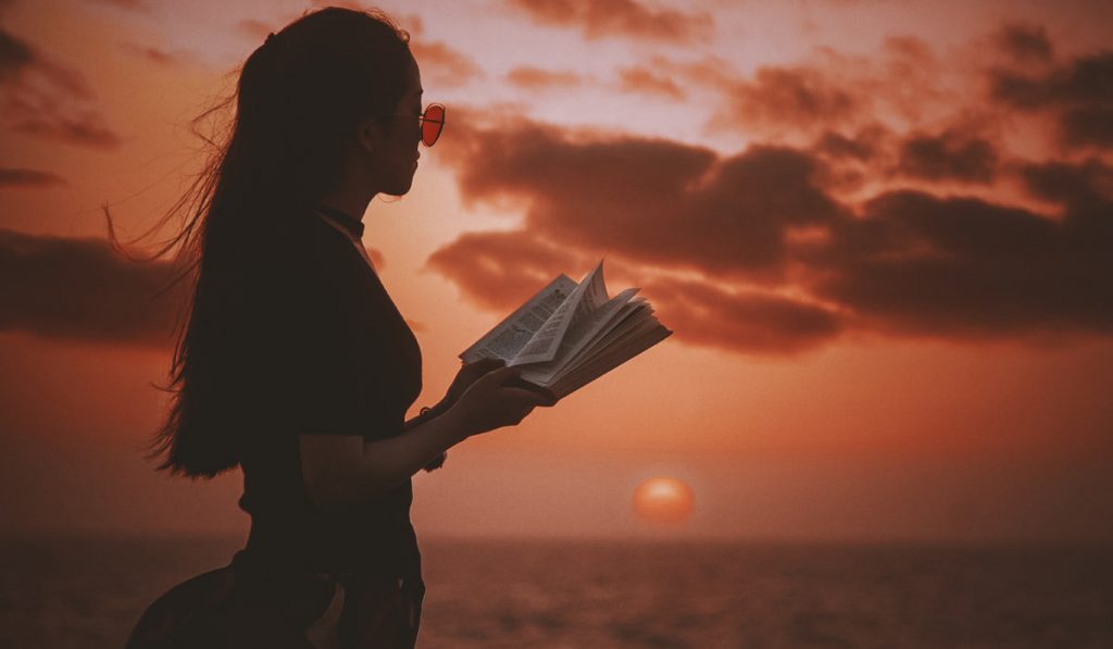 woman standing with open book, looking at sea and horizon with sunset