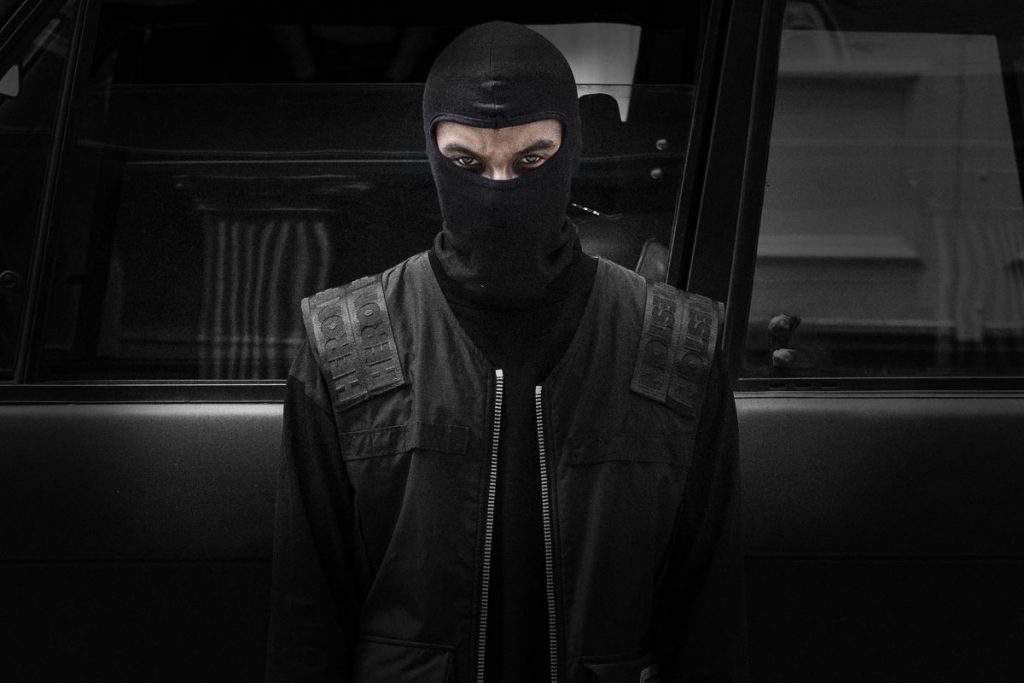 Masked man dressed in black standing by black SUV